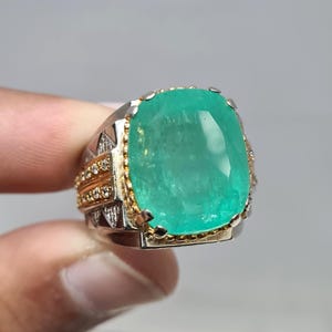 Puede incluir: Un gran anillo de piedra preciosa verde con talla esmeralda, con engaste de oro y plata. El anillo presenta una prominente piedra verde de forma rectangular, rodeada por un borde dorado y acentuada con piedras más pequeñas. La banda es plateada con detalles dorados.