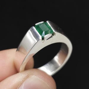 May include: A silver ring featuring a rectangular, emerald-green gemstone. The ring has a simple, modern design with a polished finish. The gemstone is set in a bezel setting, and the band is smooth and wide.