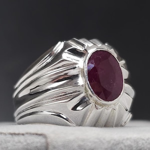 May include: A silver ring with a large, oval-cut ruby gemstone. The ring has a textured, ridged design around the band and the gemstone setting. The ruby is a deep red color, and the ring is set against a dark background.
