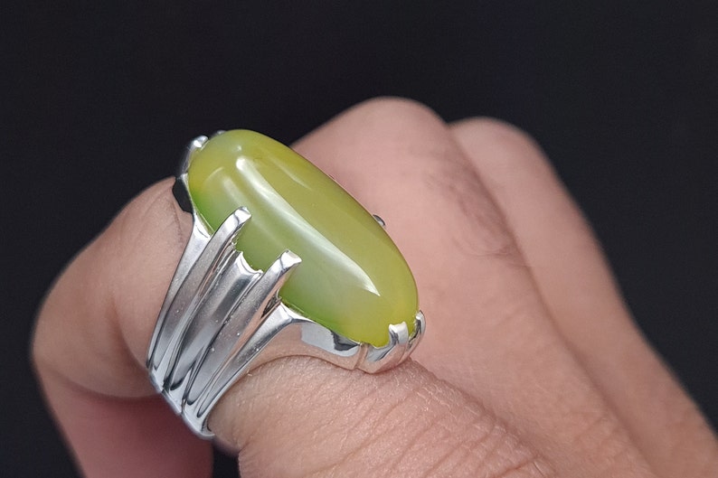 May include: A silver ring with a large, oval-shaped green gemstone. The ring has a detailed, ornate design.