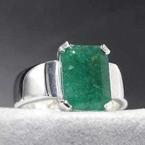 May include: A silver ring with a large, rectangular, emerald-green gemstone. The ring has a wide band and is set with four prongs. The gemstone is approximately 1.9 cm long and 1.3 cm wide.