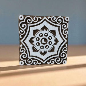 May include: Square wooden block with a floral design. The block features a white floral pattern on a dark brown background. The floral design is centered and surrounded by a decorative border. This block is likely used for printing or stamping.