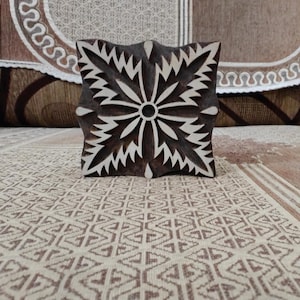 Può includere: Un blocco di legno quadrato con un disegno floreale intagliato. Il disegno presenta un cerchio centrale circondato da foglie stilizzate e radianti. Il blocco è marrone scuro con accenti bianchi e probabilmente viene utilizzato per la stampa o la timbratura.