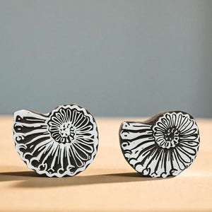 May include: Two black and white seashell-shaped decorative objects. Each has a spiral design with a central floral pattern. The objects are placed on a light-colored surface against a neutral background.