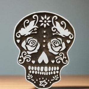 Könnte beinhalten: Ein dekorativer Zuckerschädel mit weißen Blumen- und Schmetterlingsakzenten auf dunklem braunen Hintergrund. Der Schädel hat Rosen- und Herzdetails mit einem Stern unten. Ideal für Feierlichkeiten zum Tag der Toten.