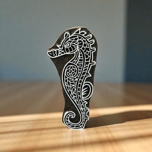 Könnte beinhalten: Ein dunkler Holzblock mit einem weißen Seepferdchen-Design. Das Seepferdchen hat komplizierte Details, darunter einen detaillierten Kopf, einen gebogenen Körper und einen Wirbelschwanz. Der Block steht auf einer Holzoberfläche.
