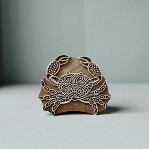 Könnte beinhalten: Ein Holzblock mit einem detaillierten Krabbenmuster. Die Krabbe ist mit symmetrischen Mustern geschnitzt, einschließlich Scheren, Körper und Beine. Der Block besteht aus hellbraunem Holz, wahrscheinlich zum Drucken oder Prägen, mit komplizierten Details.
