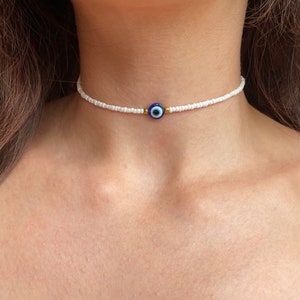 Peut inclure: Ras du cou blanc en perles avec un charme en forme d'œil bleu et doré. Le collier est composé de petites perles blanches et comporte un charme central avec un motif d'œil bleu, entouré d'accents dorés et bleus.