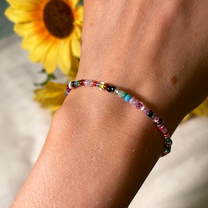 May include: A multi-coloured beaded bracelet. The bracelet is made of small, round beads in various colours, including red, blue, green, purple, black, and white. The bracelet is worn on a wrist.