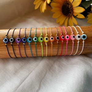 Könnte beinhalten: Ein Set aus 15 bunten Armbändern aus Schnur mit bösen Augen-Anhängern. Die Armbänder sind in einer Reihe auf einer Holzoberfläche angeordnet. Die Anhänger sind alle in verschiedenen Farben, darunter Blau, Grün, Rot und Gelb.