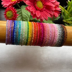 Può includere: Una pila di braccialetti colorati con perline in un motivo arcobaleno. I braccialetti sono fatti di piccole perline rotonde e sono disposti in uno schema di colore sfumato, che inizia dal viola e termina con il marrone.