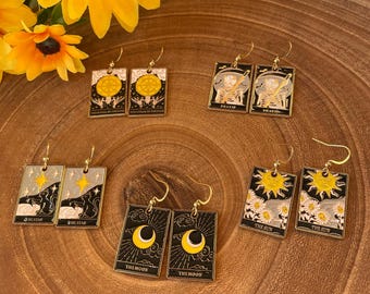 May include: A set of gold-tone earrings featuring tarot card designs. The cards include The Wheel of Fortune, Death, The Star, The Sun, and The Moon. Each card is outlined in black and has a gold-tone finish.