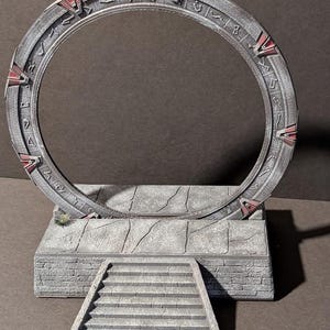 Puede incluir: Modelo detallado de un Stargate, con un gran anillo circular gris con intrincadas tallas y detalles rojos. El anillo se asienta sobre una plataforma de piedra con una escalera que conduce a él, creando una escena de ciencia ficción.
