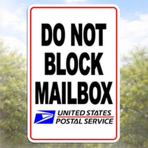 May include: A rectangular white sign with a red border displays the bold text "DO NOT BLOCK MAILBOX." Below, the United States Postal Service logo and text are visible. The background is a blurred view of trees and sky.