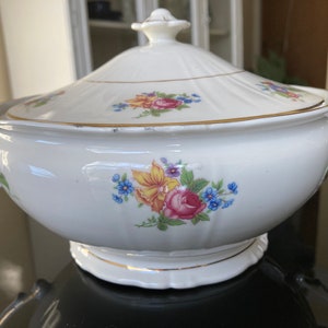 May include: A white porcelain serving bowl with a lid, adorned with floral patterns in pink, yellow, and blue. The bowl has gold trim and handles, and sits on a scalloped base. A classic piece for serving soups or stews.