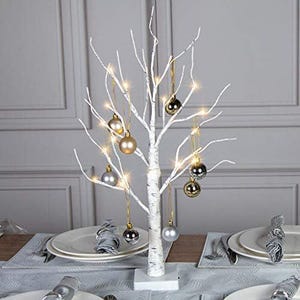 May include: A white birch tree with a white trunk and branches decorated with silver and gold baubles. The tree is lit with white fairy lights and is sitting on a white base.