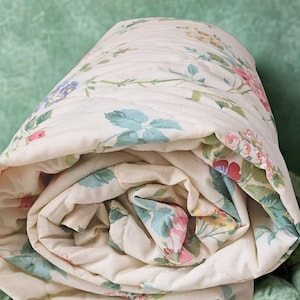 May include: Rolled floral quilt with a cream base and a pattern of pink, blue, and yellow flowers and green leaves. The quilt is made of a soft, quilted material and is resting on a green textured surface.