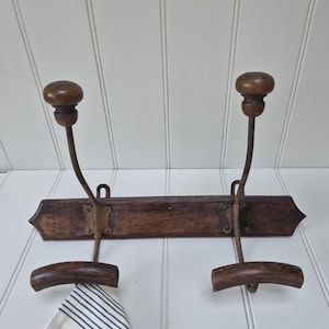 May include: A wall-mounted coat rack featuring a dark brown wooden rectangular base with pointed ends. It has two metal hooks, each topped with a round wooden knob and a curved wooden bar at the bottom. A white cloth with thin black stripes hangs from one hook.
