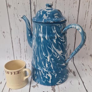 May include: A blue and white enamelware coffee pot with a matching lid and handle. Beside it is a beige mug with the word "OFFEE" printed on it. The coffee pot has a classic, vintage design.