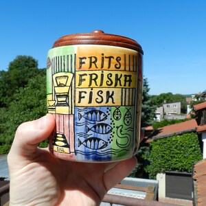 Op de afbeelding: Een cilindervormige keramische pot met een donkerbruin houten deksel. De buitenkant is voorzien van handgeschilderde illustraties van vissen en keukenmotieven in geel, blauw en groen, met de zwarte tekst FRITS FRISKA FISK en SILL erop.