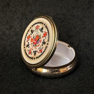 Op de afbeelding: Een open, ovale pillendoos met een bloemmotief op het deksel. Het deksel heeft een crèmekleurige achtergrond met een kleurrijk bloemmotief en een zwarte rand. De doos is zilverkleurig met een witte binnenkant.