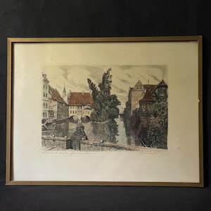 Pode incluir: Uma gravura emoldurada retratando uma cena de canal em Nuremberga, Alemanha, com edifícios históricos, uma ponte de pedra e árvores. A obra é apresentada em tons suaves de castanho, verde e cinzento, com uma assinatura e um título manuscrito.