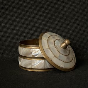 May include: A decorative box with a lid, featuring a mother-of-pearl inlay design. The box has brass trim and a brass knob on the lid. The box is open, revealing the interior. The box is on a black surface.