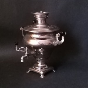 May include: A silver-colored samovar, a traditional Russian tea urn, stands against a dark background. The samovar features a bulbous body, a spigot, and a chimney-like top. It has two handles and sits on a square base.
