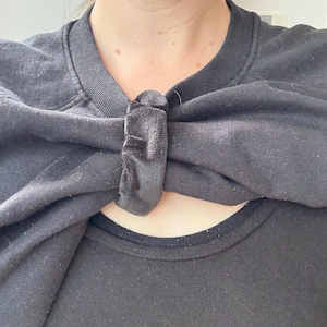 May include: A black sweatshirt with a bow-like detail at the neckline. The bow is made of the same material as the sweatshirt. The neckline of a black t-shirt is visible underneath.