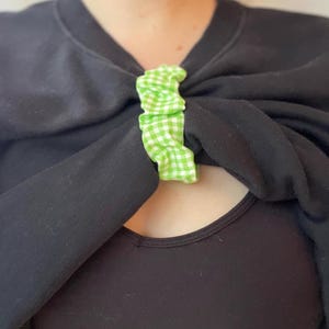 May include: A green and white gingham fabric hair tie is used to hold a black shirt together at the neckline.