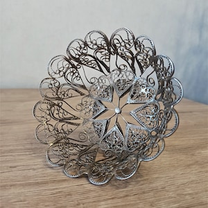 May include: A silver filigree bowl with an intricate, openwork design. The bowl features a floral pattern with heart-shaped elements and a central starburst. The delicate metalwork creates a lacy effect, showcasing the craftsmanship of the piece.