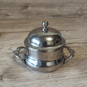 Op de afbeelding: Een zilverkleurige suikerpot met deksel en sierlijke handvatten. De kom heeft een afgeronde vorm en een gewelfd deksel met een kleine knop. De handvatten zijn gebogen en gedetailleerd. De suikerpot staat op een houten oppervlak.