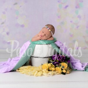 May include: A sleeping baby wearing a flower headband lies in a white wooden bucket with a green and purple fabric draped over the sides. Yellow and purple flowers are in front of the bucket.