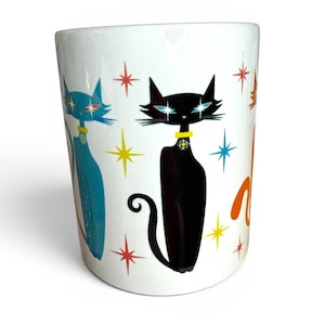 May include: White ceramic mug featuring mid-century modern cat illustrations in blue, black, and orange. The cats wear yellow collars and are surrounded by starburst designs in various colors. A retro-style design.