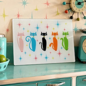 May include: A rectangular glass cutting board featuring a graphic illustration of five stylized cats in pink, blue, black, orange, and green. The cats are arranged in a row against a white background decorated with scattered multi-colored starbursts.