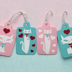 May include: Four decorative gift tags with a cat design. Two tags are pink, and two are teal. Each tag features a white cat illustration with a red heart and a silver string for hanging. The cat has a bow tie.