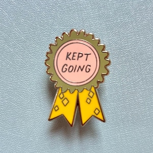 May include: A gold-colored enamel pin with a green and pink circle that says "KEPT GOING" and a yellow ribbon with diamond-shaped patterns.