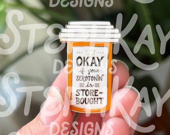 May include: A gold and orange enamel pin shaped like a pill bottle with the text "It's okay if your serotonin is store-bought".