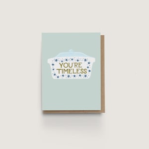 May include: A greeting card with a light blue background and a brown border. The card features a white casserole dish illustration with blue snowflake accents and the gold text "YOU'RE TIMELESS."