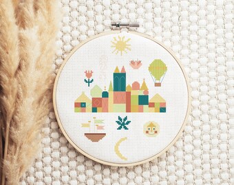 May include: A circular wooden embroidery hoop containing white fabric with a cross-stitch design. The pattern features a colorful stylized cityscape with geometric buildings, a yellow sun, a hot air balloon, a boat, a moon, and a smiling face.