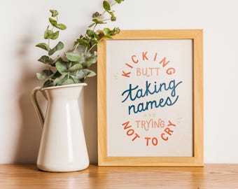 May include: A framed print with a motivational quote in blue and orange text on a beige background. The quote reads "Kicking Butt Taking Names And Trying Not To Cry". The frame is made of light wood and is sitting on a light wood surface.