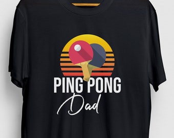 Könnte beinhalten: Schwarzes T-Shirt mit einem Retro-Sonnenuntergang-Motiv und zwei Tischtennis-Schlägern. Der Text "PING PONG Dad" ist unter dem Motiv aufgedruckt.