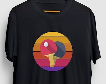 Puede incluir: Camiseta negra con un gráfico retro de dos palas de ping pong y una pelota en un círculo con un fondo de puesta de sol.
