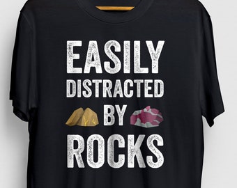 Puede incluir: Camiseta negra con texto blanco que dice "Easily Distracted by Rocks" con dos pequeñas ilustraciones de rocas.