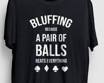May include: Black t-shirt with white text that reads "Bluffing because a pair of balls beats everything".  The text is surrounded by a circle and there are four playing card suit symbols below the text.