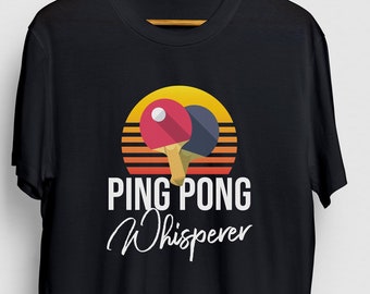 Puede incluir: Camiseta negra con un gráfico de puesta de sol retro y dos palas de ping pong. El texto "PING PONG Whisperer" está impreso en blanco debajo del gráfico.