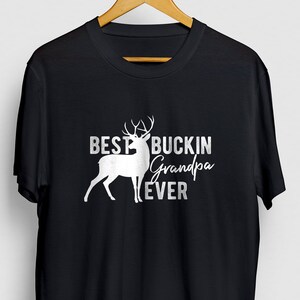 K&ouml;nnte beinhalten: Schwarzes T-Shirt mit einer wei&szlig;en Silhouette eines Hirsches und dem Text "Best Buckin' Grandpa Ever".