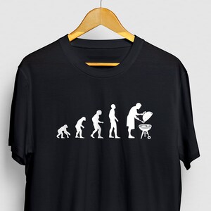 May include: Black t-shirt with a white graphic of the evolution of man, ending with a person grilling. The graphic shows a series of stick figures, starting with a primitive ape and ending with a person standing at a grill.
