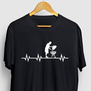 May include: Black t-shirt featuring a white graphic of a person barbecuing with a heartbeat line above it.