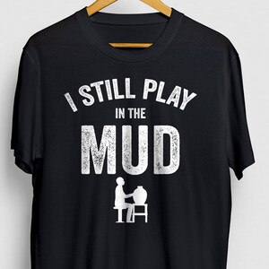 May include: Black t-shirt with white text that reads "I still play in the mud" and a silhouette of a person using a pottery wheel.